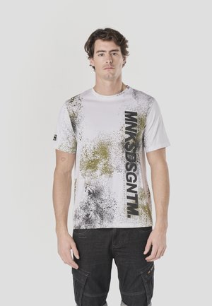 Wit T-shirt met een spatterdesign in zwart en olijfgroen, met de tekst "MNKSDSGNTM" in vet zwart verticaal langs één zijde.