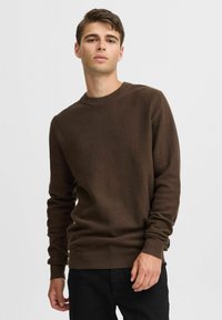 Brun genser i strikk med rund hals, teksturert mønster og ribbede mansjetter. Brukt uformelt med mørke jeans, som viser en avslappet passform.
