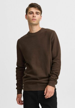 Brun genser i strikk med rund hals, teksturert mønster og ribbede mansjetter. Brukt uformelt med mørke jeans, som viser en avslappet passform.
