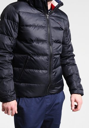 Muž v černé prošívané péřové bundě se zapínáním na přední zip a kapsami po stranách, v kombinaci s tmavě modrými kalhotami na jednobarevném pozadí.