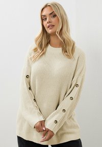 Jasnobeżowy sweter z dzianiny o luźnym kroju, z dużymi guzikami wzdłuż rękawów i we wzór strukturalny. Miękka i przytulna tkanina.