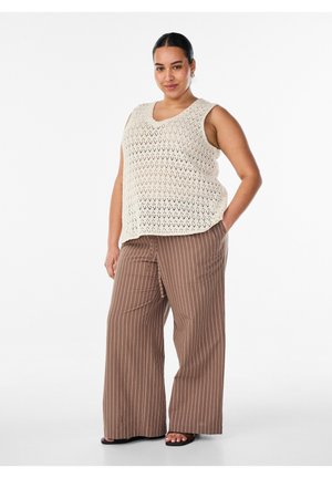 Mujer con cabello oscuro, con un top de punto sin mangas beige y pantalones marrones de pierna ancha con rayas blancas, de pie con una mano en el bolsillo.
