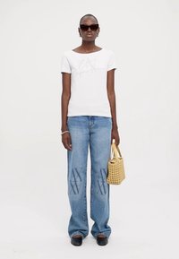 Camiseta de algodón blanca con 'EA' impreso en plata, combinada con unos pantalones vaqueros de denim azul claro con detalles gráficos, y sosteniendo un bolso tejido amarillo.