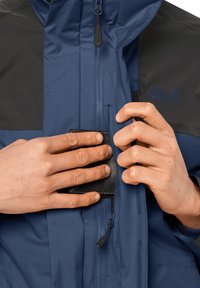 Niebieska i czarna wodoodporna kurtka z zamkami błyskawicznymi, kieszeniami i logo. Ręce dostosowują mały czarny dodatek w kieszeni na klatce piersiowej.