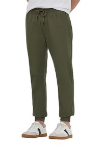 Pantaloni elasticizzati di colore verde oliva con vita regolabile, polsini affusolati e tasche laterali, abbinati a sneakers bianche con dettagli neri.