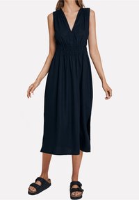 Robe sans manches bleu marine avec un décolleté en V profond et des épaules froncées. Comprend une taille élastique et une fente latérale. Associée à des sandales noires à enfiler.