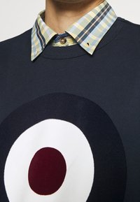 Granatowa bluza z dużym, okrągłym nadrukiem w białym, czerwonym i ciemnoniebieskim kolorze. Posiada kołnierz w kratę w odcieniach zieleni, żółci i niebieskiego.