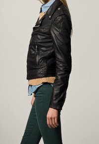 Veste de motard en cuir noir avec des fermetures éclair, des épaulettes et des patches texturés sur les épaules, portée sur un pull beige et une chemise bleue.