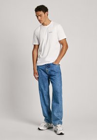 Bílý bavlněný tričko s malým modrým logem, spárované s volně střiženými modrými džíny a šedými teniskami. Neformální outfit s jednoduchým designem.