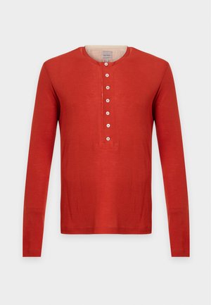 Homme portant un t-shirt henley à manches longues orange, un jean large bleu clair et des baskets blanches, debout devant un fond uni.