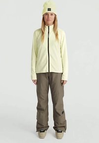 Jasnożółta polarowa kurtka z zamkiem z przodu, w połączeniu z luźnymi oliwkowymi spodniami i pasującą czapką. Obuwie w kolorze beżowym z czarnymi akcentami.
