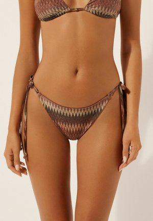Femme portant un bikini marron à motif zigzag avec bas à nouer sur les côtés, visible de la moitié du torse jusqu'aux cuisses, sur un fond uni.