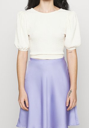 Kobieta w białym teksturowanym krótki top z bufkami na rękawach i wysokim stanem lawendowej satynowej spódnicy, pokazująca tatuaże na ramionach.