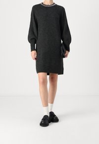 Robe en maille grise avec manches longues bouffantes, col rond souligné d'une bordure métallique, associée à des chaussures noires sans lacets et des chaussettes blanches.