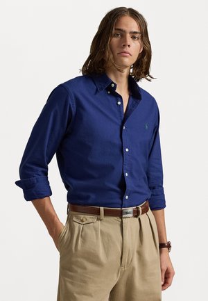 Cămașă bleumarin, cu nasturi, din bumbac, cu mâneci suflate, având un mic logo verde pe pieptul stâng. Asortată cu pantaloni beige plisati.
