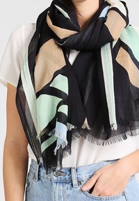 Écharpe à motifs noirs, beiges et menthe, fabriquée en tissu léger avec des bords frangés. Drapée librement autour du cou.