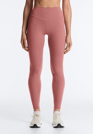 Femme portant des leggings taille haute rose poussiére et des baskets blanches, debout devant un fond clair uni.