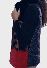 Vesta z umělé kožešiny s černým prošívaným zadním dílem, leopardím potiskem na přední straně a červeným kožíškovým lemem. Detail se zipem u krku zdůrazňuje design.