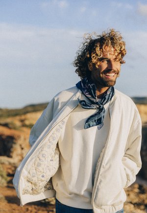 Mężczyzna o kręconych włosach uśmiechający się na zewnątrz, ubrany w biały otwarty płaszcz ukazujący wewnętrzną podszewkę z wzorem słonecznym oraz ciemnoniebieski szalik wokół szyi.