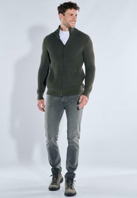 Olive green, ribbed zip-up sweater worn over a white t-shirt, paired with light gray jeans and gray hiking boots. Simple design, relaxed fit.