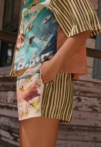 Personne portant un short à rayures vertes avec un imprimé aquarelle plage, main dans la poche, associé à une chemise présentant des surfeurs et des scènes océaniques.