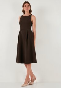 Kobieta ubrana w bezrękawową, ciemnobrązową midi sukienkę z zakładkami oraz beżowe, szpiczaste, odkryte czółenka na tle białym.