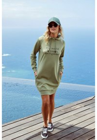 Olivenfarbene Kapuzenkleid mit dem Text "Die Welt gehört dir zu erkunden", gerippte Bündchen und Saum, kombiniert mit schwarzen Sneakers und einer passenden Kappe.