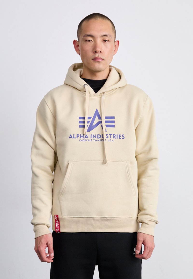 Šviesiai rudas džinsinis džemperis, pagamintas iš minkštos medžiagos, su priekinėmis kišenėmis, reguliuojama kapucė ir dideliu mėlynu logotipu "ALPHA INDUSTRIES". Tekstūruoti rankogaliai ir apačia.
