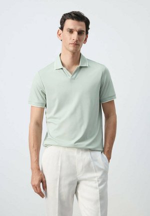 Hombre con cabello oscuro, vestido con una camisa polo verde claro y pantalones blancos, de pie con una mano en el bolsillo contra un fondo liso.
