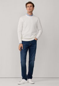 Witte, lichte trui met V-hals, gecombineerd met blauwe denim jeans en witte sneakers. Gestreepte kraag zichtbaar onder de trui.