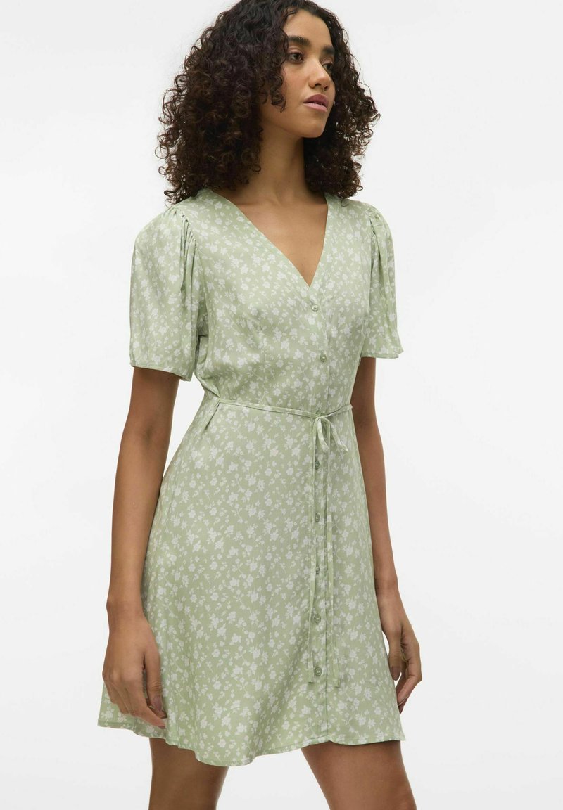 Robe florale vert clair avec des manches courtes à volants, un décolleté en V et une fermeture à boutons. Comprend une ceinture à nouer et une silhouette fluide.
