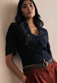 Blusa negra de manga corta con botones y cuello, combinada con pantalones de talle alto color óxido y un cinturón negro tejido con hebilla circular.