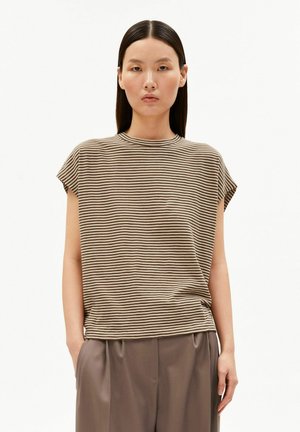 Frau mit glatten schwarzen Haaren, die ein beige-braun gestreiftes kurzärmeliges Shirt und braune Hochbundhosen trägt, steht mit den Händen in den Taschen.