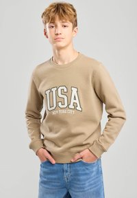 Beige sweatshirt featuring "USA NEW YORK CITY" printed in bold letters, round neckline, and ribbed cuffs. Paired with blue jeans.