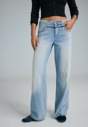 Donna che indossa jeans blu sbiaditi a vita alta con gamba larga e doppio bottone in vita, con ballerine nere, una mano in tasca.