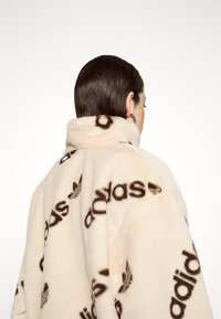 Persona con capelli scuri voltata di spalle, che indossa una giacca in pile color crema con stampa integrale del logo Adidas nero e un orecchino a cerchio.