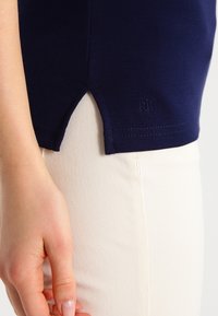 Detailní záběr na ruku vedle lemu tmavě modré košile s bočním rozparkem, krémových kalhot a vyšívaného loga "RL" poblíž lemu.