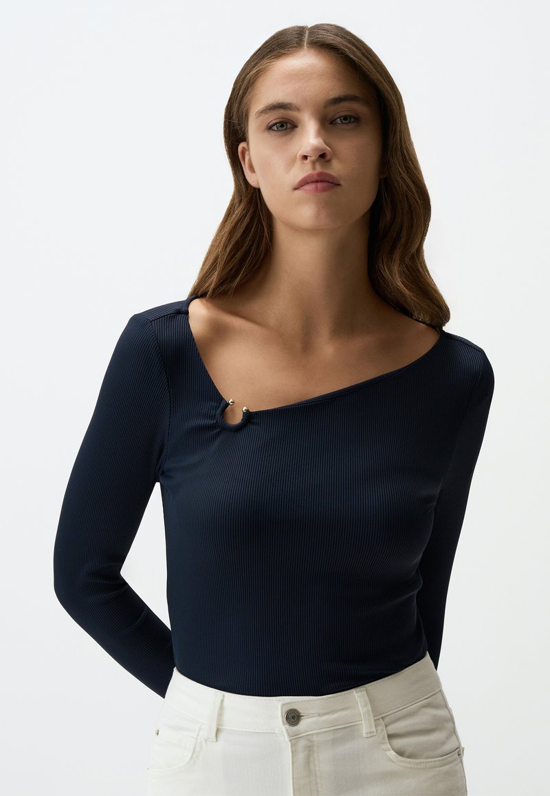 Maglietta a maniche lunghe a coste blu navy con scollatura asimmetrica, decorata con un dettaglio metallico sulla spalla, abbinata a pantaloni bianchi.