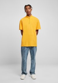 Gul kortermet polo skjorte med høy krage og glidelås, sammen med lyseblå vaskede jeans og hvite joggesko.