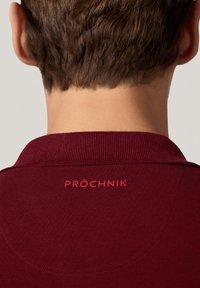 Bordowa koszulka polo z teksturowanej tkaniny. Posiada prążkowany kołnierzyk oraz czerwoną wyszywaną naszywkę "PRÓCHNIK" na plecach.