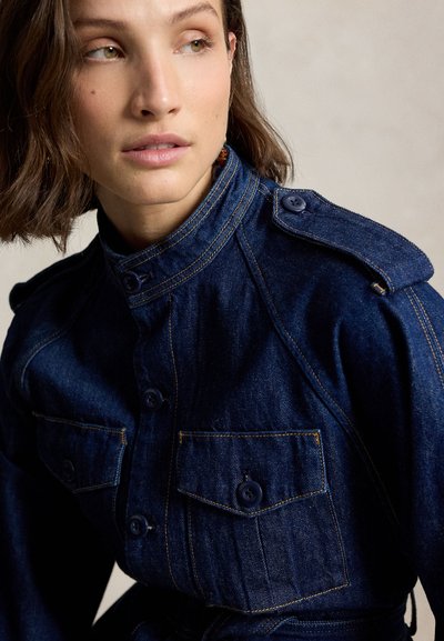 Ženska s kratkimi rjavimi lasmi, oblečena v temno modro jeans jakno z zaponkami na prsnih žepih in škornji, gleda vstran na enobarvni ozadju.