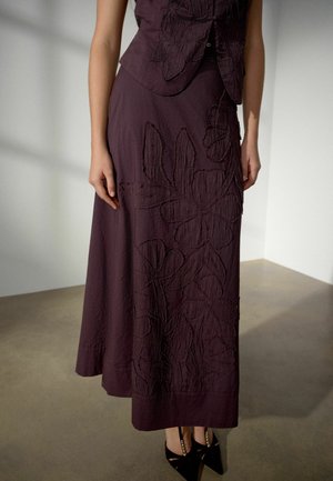 Sieviete valkā tumši violetu svārku ar lielu ziedu izšuvumu, saskaņotu ar melniem punktveida augstpapēžiem ar zelta rotājumiem.