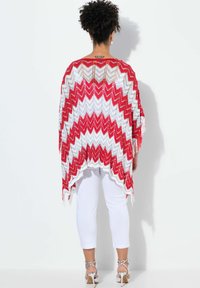 Poncho a zigzag rosso e bianco con dettagli a frange, realizzato in un materiale knit leggero, indossato sopra pantaloni bianchi a sigaretta e tacchi con cinturini.