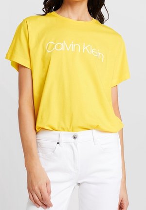 Person trägt ein gelbes Calvin Klein T-Shirt, das in weiße Hosen gesteckt ist, eine Hand ruht nahe der Hüfte.