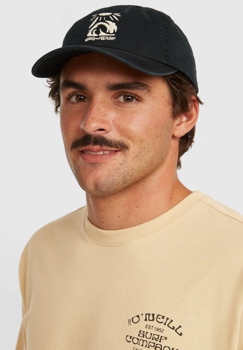 Gorra negra con un diseño gráfico blanco en la parte frontal, llevada por una persona con una sudadera beige que presenta texto impreso y un cuello redondeado.