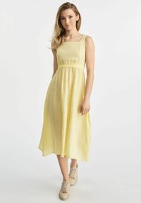 Żółta sukienka do kolan z kwadratowym dekoltem, zebranym w talii i lekką teksturą materiału. Połączona z beżowymi butami na sznurowadła.