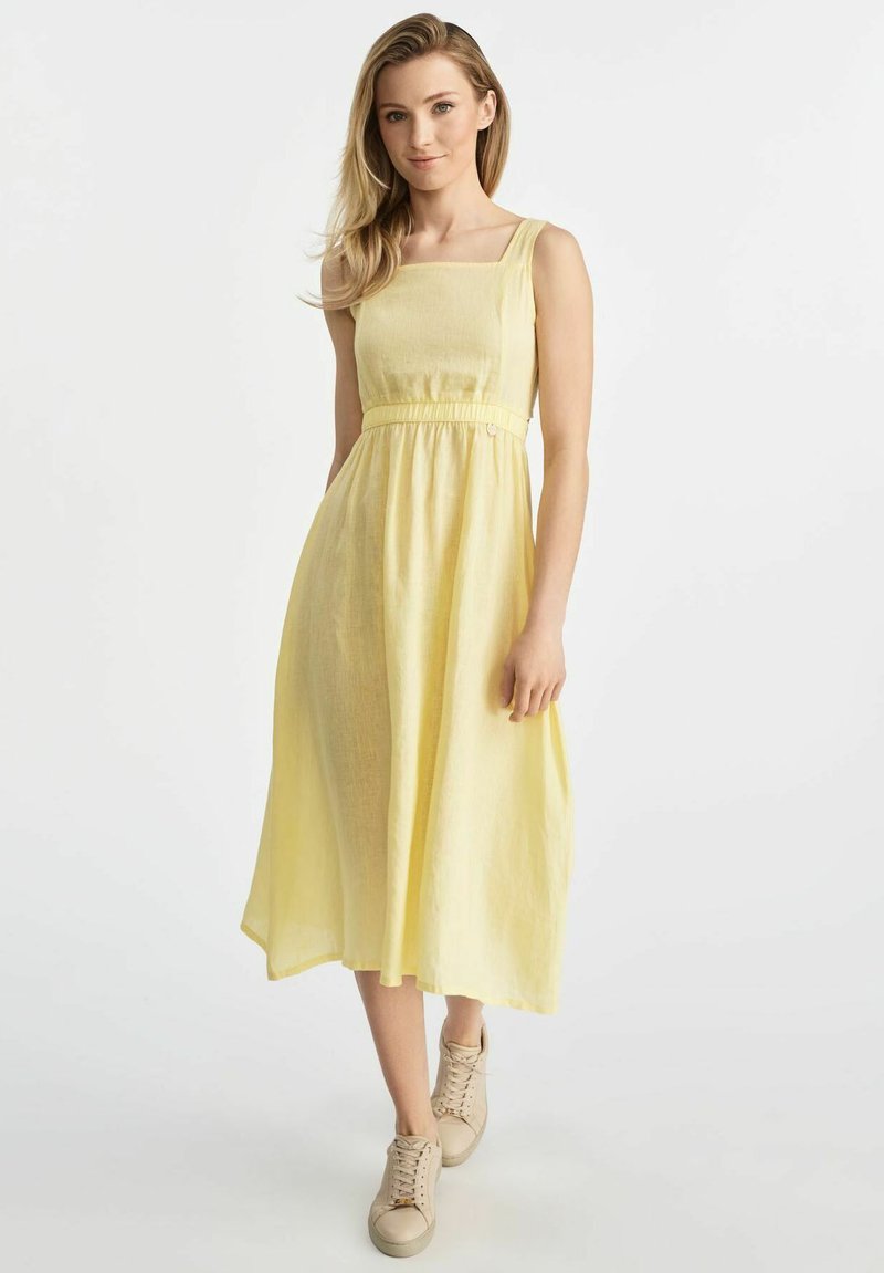 Żółta sukienka do kolan z kwadratowym dekoltem, zebranym w talii i lekką teksturą materiału. Połączona z beżowymi butami na sznurowadła.
