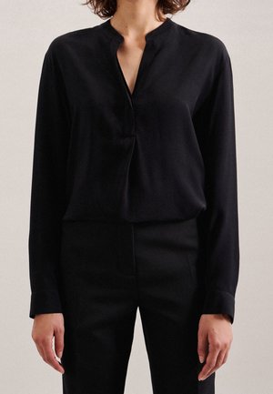 Vrouw die een zwarte blouse met lange mouwen en een inkeping in de hals draagt, en een zwarte broek, staand tegen een effen lichte achtergrond.