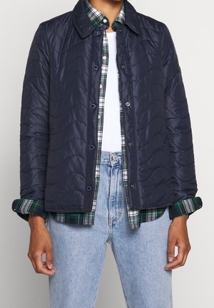 Osoba ubrana warstwowo, ma na sobie granatową pikowaną kurtkę na zielono-białej koszuli w kratę oraz jasnoniebieskie dżinsy.