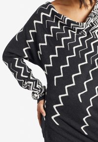 Kobieta w czarnym swetrze z białymi wzorami w zygzak i szerokim dekoltem, opierająca rękę na biodrze na białym tle.